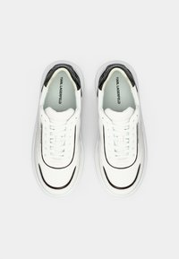 Λευκά αθλητικά παπούτσια με κομψό σχεδιασμό, που διαθέτουν γυαλιστερή μαύρη λεπτομέρεια, στρογγυλή μύτη, επίπεδα κορδόνια και λογότυπο στο πλάι.