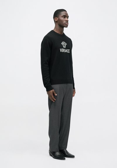 Černý svetr s vyšitým logem "VERSACE" a emblémem Medúzy, doplněný o šedé pouzdrové kalhoty a černé boty, stojící na jednoduchém pozadí.