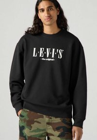 Felpa nera con grafica "LEVI'S the original" in bianco, dal fit morbido e polsini a costine. Abbinata a pantaloni con stampa camouflage.