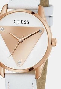 Roosa kuldne kell valge näo, kolmnurkse disaini ja valge nahkrihmaga. Kella omadused hõlmavad minimalistlikke tundi ja minuti näpse hõbedaste aktsentidega.