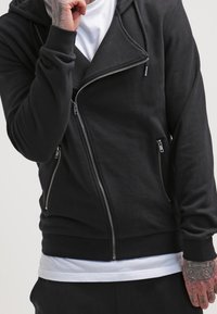 Sudadera con cremallera negra con un diseño de motocicleta, hecha de una tela suave. Cuenta con una doble cremallera y bolsillos frontales. Superpuesta a una camiseta blanca.