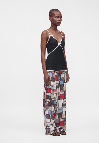 Femme aux cheveux courts portant un top caraco noir avec des bordures blanches et un pantalon large coloré avec un imprimé de dos de livre, debout devant un fond blanc.