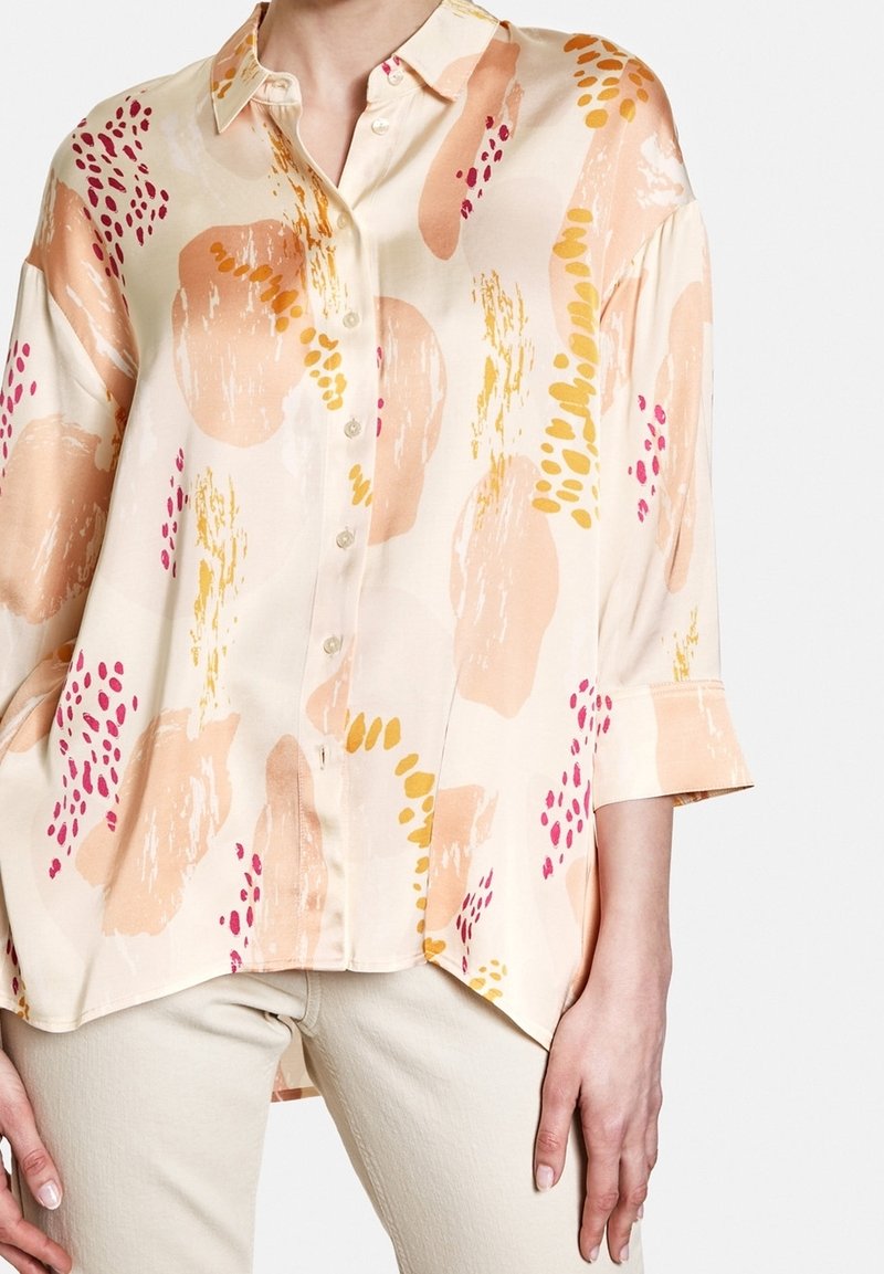 Chemise ample en crème à boutons, ornée de motifs abstraits en coups de pinceau pêche, jaune et rose, portée avec un pantalon beige clair.