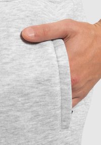 Helle graue Jogginghose mit einer Seitentasche, aus einem weichen, strukturierten Stoff. Eine Hand ist teilweise in der Tasche sichtbar.