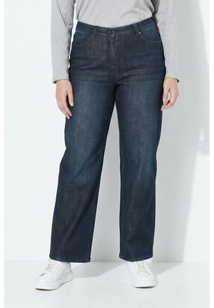 Person trägt dunkelblaue Jeans mit geradem Bein, weiße Sneakers und ein hellgraues Langarmshirt und steht vor einem weißen Hintergrund.