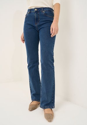 Kvinde iført blå jeans med lige ben og beige vævede flade sko, stående mod en ensfarvet lys baggrund.