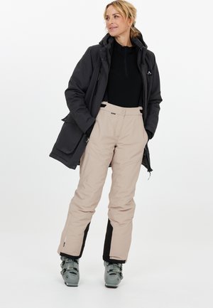 Skidräkt med en svart dragkedjetröja, en mörk jacka, beige byxor med svarta detaljer och grå skidstövlar, designad för vintersporter.