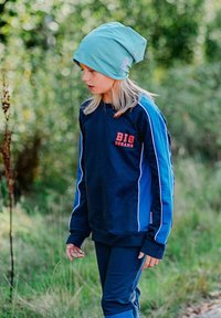 Sinili ja tumesinis tracksuit valgete triipudega ja tekstiga "SUURED UNED". Pehme kangast beanie helesinises. Mudel seisab õues, rohurikas taust.