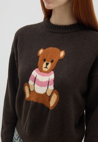 Brauner Strickpullover mit Bärenmotiv, pinken und weißen Streifen, rundem Ausschnitt und weichem Material.