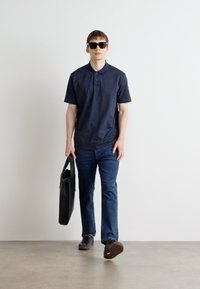 Camisa polo azul-marinho, jeans de ganga e óculos de sol pretos, a segurar uma mala preta; ajuste casual, tecido suave e design clássico.