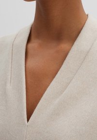 Top beige chiaro a scollo a V realizzato in un tessuto morbido e testurizzato, con una sottile lucentezza e linee pulite, caratterizzato da un design minimalista senza decorazioni.