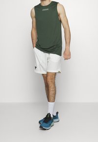 Zielona bezrękawnik sportowy, białe szorty i niebieskie buty sportowe z czarnymi akcentami. Buty mają siateczkową górę i wyściełaną podeszwę.