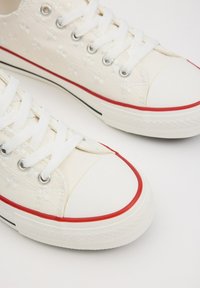 Zapatillas de lona blancas con bordado floral, suela de goma roja, puntera reforzada y cordones blancos. Ribete negro a lo largo de la suela.