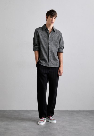 Camisa de botones gris con patrones, pantalones negros texturizados y zapatos tipo slip-on con un colorido patrón en zigzag. El modelo está de pie con las manos en los bolsillos.