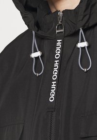Giacca con cappuccio nera con zip frontale, coulisse bianche e dettagli con logo stampato. Presenta una texture liscia e tasche laterali.