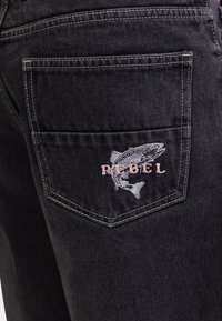 Sorte denim jeans har en baglomme med hvid syning og et broderet design af en fisk med ordet "REBEL" i pink.