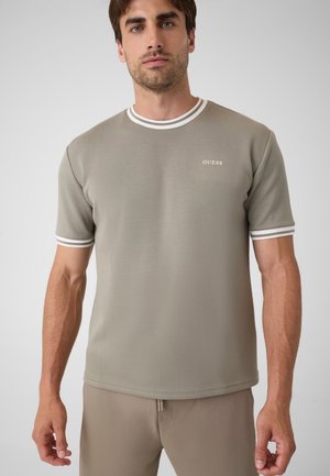 Mann trägt ein taupefarbenes Kurzarmhemd mit weiß gestreiftem Kragen und Manschetten und kleinem "GUESS" Logo auf der Brust.