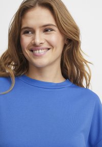 Sweatshirt in cotone blu con collo rotondo a costine. Presenta una texture liscia e una vestibilità rilassata, mostrando una parte del sorriso di chi lo indossa.