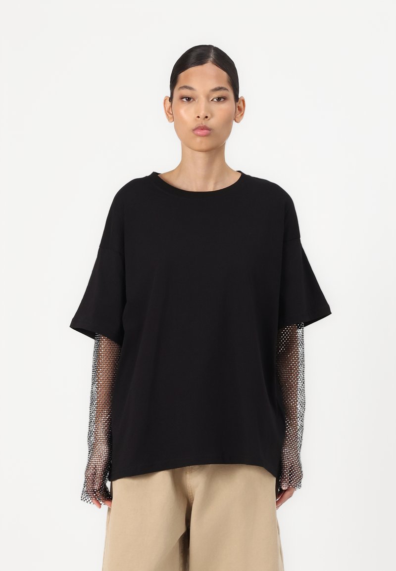 Svart oversized t-skjorte med korte ermer; mesh lange ermer med rutemønster. Stoffet ser mykt ut; avslappet passform. Naturlig fargede bukser.