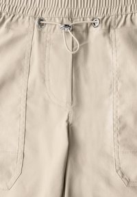 Pantaloni beige con vita elastica, dotati di cordoncino e tasche frontali. Texture liscia e dettagli delle cuciture sartoriali.