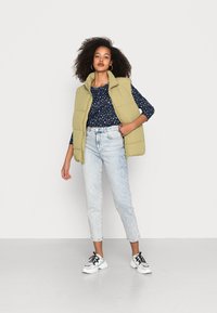 Colete verde acolchoado sobre uma camisa azul com manchas, combinado com jeans de lavagem clara e ténis preto e branco. Foco na textura e nas sobreposições.