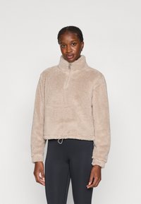 Veste polaire en peluche, beige, avec un col haut et une fermeture éclair d'un quart de longueur. Comprend un cordon de serrage ajustable à l'ourlet.