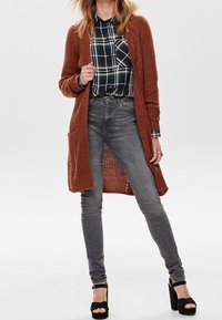 Cardigan de punto marrón oxidado sobre una camisa de cuadros abotonada, combinado con jeans skinny grises y tacones de plataforma negros. El cardigan cuenta con bolsillos abiertos.