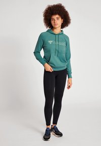 Tealfärgad hoodie med framficka och logotyp, i kombination med svarta leggings. Modellen bär svarta och blå sportskor. Texturerat tyg.