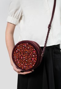 Borsa circolare in pelle bordeaux con ricami floreali intricati in rosso e arancione. Ha una tracolla intrecciata e metalleria dorata.