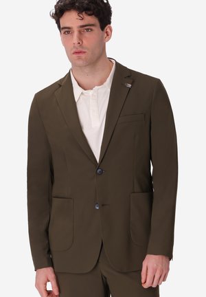 Uomo che indossa una giacca da completo verde oliva sopra una polo bianca, in piedi davanti a uno sfondo chiaro e uniforme con espressione neutra.