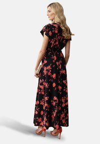 Schwarzes Maxikleid mit blühendem Muster, kurzen schwingenden Ärmeln, geraffter Taille und roten Blumenmustern; kombiniert mit roten High Heels und einer metallischen Clutch.