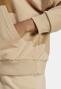 Mano appoggiata all'interno della tasca anteriore di una felpa beige con polsini e bordo a coste, pantaloni beige abbinati visibili.