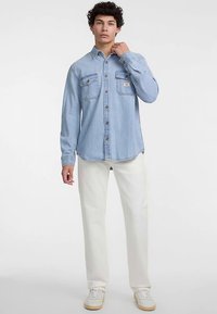 Lyseblå denimskjorte med knaplukning foran, brystlommer og kontrastsyninger, kombineret med hvide bukser og hvide sneakers.