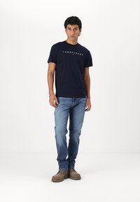 Marineblå bomull T-skjorte med "TOMMY JEANS"-logo, lys blå denim jeans og beige sko. Enkel design, korte ermer, avslappet passform.