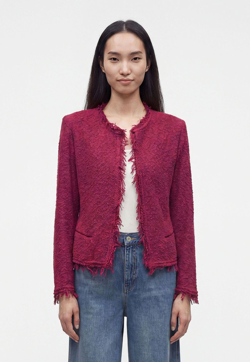 Femme aux longs cheveux foncés portant une veste rouge texturée aux bords effilochés, un haut blanc et un jean bleu, debout devant un fond uni.
