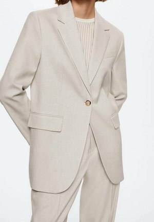 Persona che indossa un abito beige chiaro su misura con un singolo bottone dorato e tasche con patta sopra un top crema a coste.