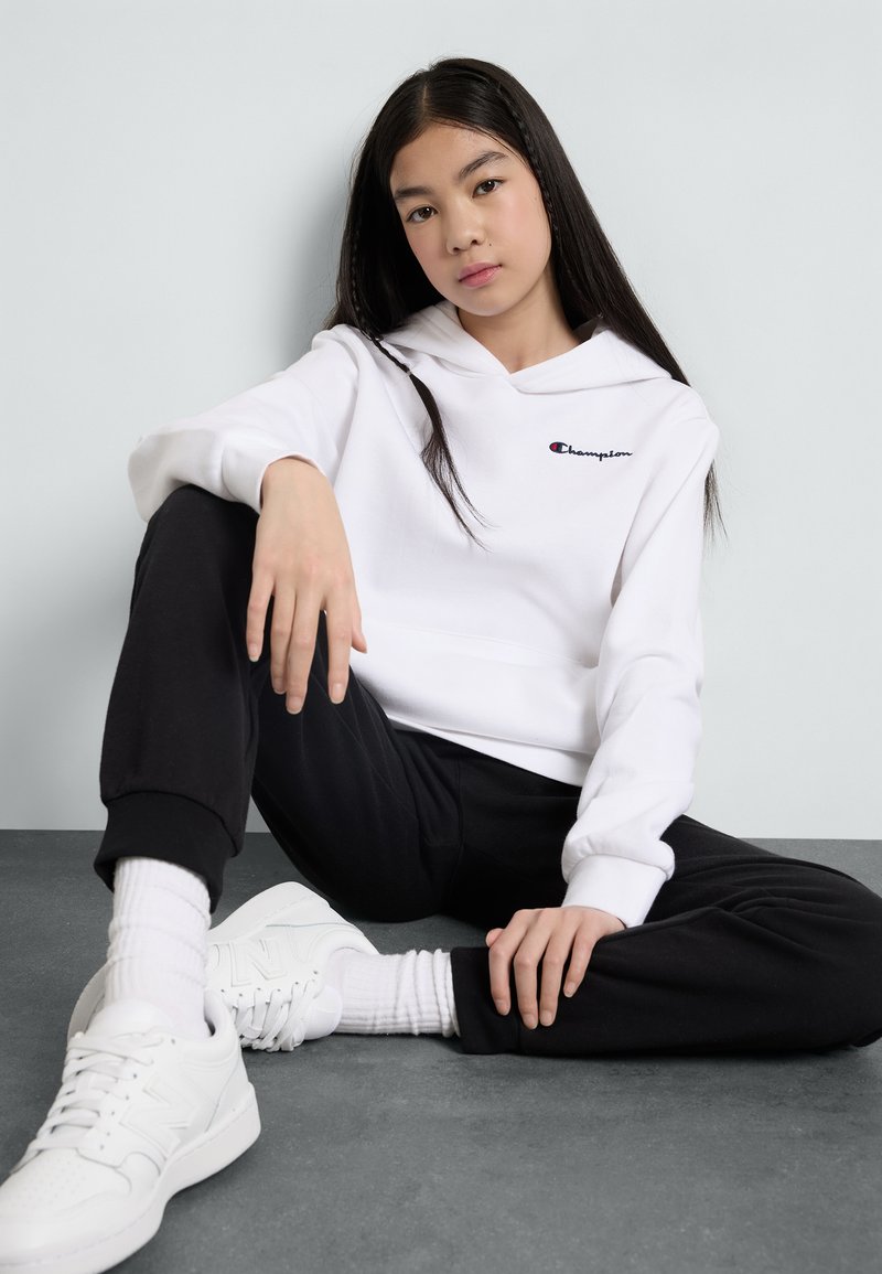 Felpa bianca con cappuccio e un piccolo logo rosso e blu, abbinata a pantaloni jogger neri e scarpe da ginnastica bianche. Modello seduto su una superficie grigia.