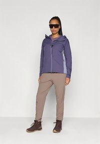 Fioletowa lekka kurtka z zamkiem błyskawicznym, szarymi akcentami i kieszeniami; w zestawieniu z beżowymi spodniami i brązowymi butami trekkingowymi. Gładka tekstura.