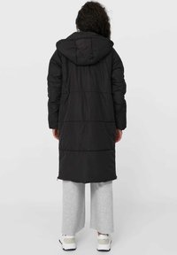 Lang sort dunjakke med hætte, der har quiltede sektioner, en afslappet silhuet og en glat overflade. Båret over lyse grå sweatpants og sko.