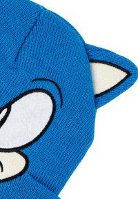 Blauwe gebreide muts met grote witte ogen en zwarte omlijningen, voorzien van een witte en zwarte dierenoor aan de rechterkant.