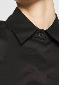 Maglietta nera con colletto classico, dotata di chiusura a bottoni sul collo e texture del tessuto liscia e opaca. Dettagli di design minimalista.