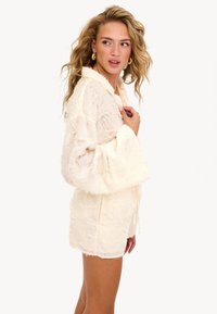 Femme jeune avec des cheveux blonds ondulés, portant une veste texturée blanc cassé et un short assorti, regardant sur le côté contre un fond blanc uni.