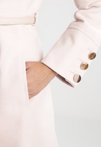 Manteau rose clair en tissu texturé, avec une taille ceinturée, des poches diagonales et trois boutons métalliques ronds au niveau des poignets.