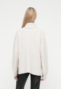 Maglione oversize color beige chiaro con finitura testurizzata, colletto alto e design con spalla cadente. L'orlo è leggermente più lungo sul retro.