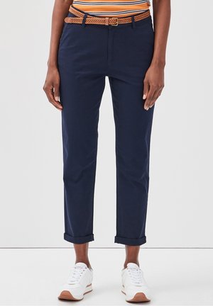 Personne portant un pantalon bleu marine retroussé, des baskets blanches, une chemise rayée orange, et une ceinture tressée marron clair, debout devant un fond uni.