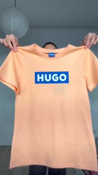 Une personne tient un t-shirt couleur pêche avec un rectangle bleu et le texte blanc « HUGO » sur le devant, dans un cadre intérieur simple.
