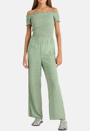 Jumpsuit floral vert avec décolleté asymétrique, corsage smocké, pantalon large et tissu léger. Des sandales argentées complètent le look.