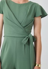 Grön jumpsuit med korta ärmar, omslagdesign vid midjan, knytband som accent, tillverkad av ett slätt tyg med en subtil lyster. Guldarmband i manschett.