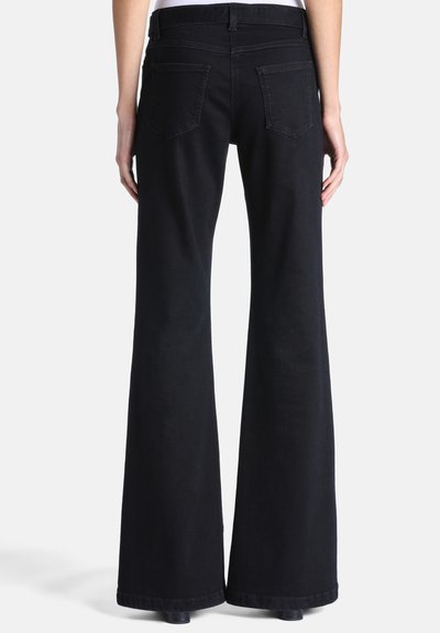 Personne portant un jean noir à jambes larges, debout, dos à la caméra, sur un fond uni.