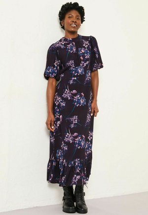 Floral Maxi-Kleid in Dunkelviolett mit Puffärmeln, gewelltem Saum und Blumenmuster in Blau/Rosa, kombiniert mit schwarzen Knöchelschuhen.
